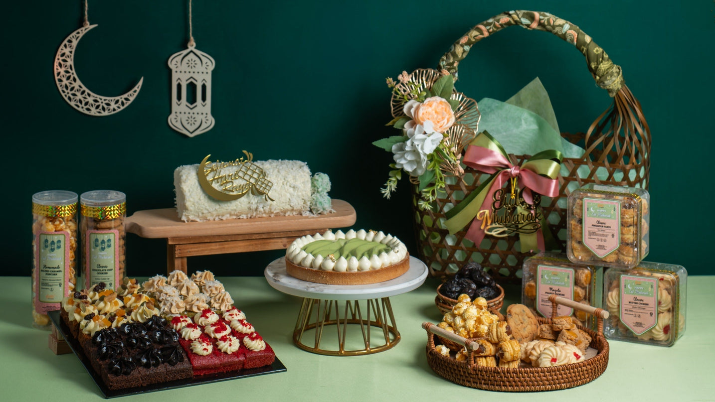 Raya hamper display with woven basket, cookie tubes and cakes against a green backdrop with a crescent lantern — Elevete Patisserie