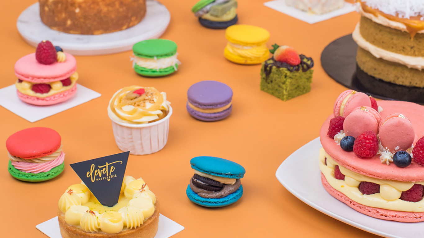 Macarons, fruit tarts and a layer cake arranged on an orange backdrop — Elevete Patisserie KL