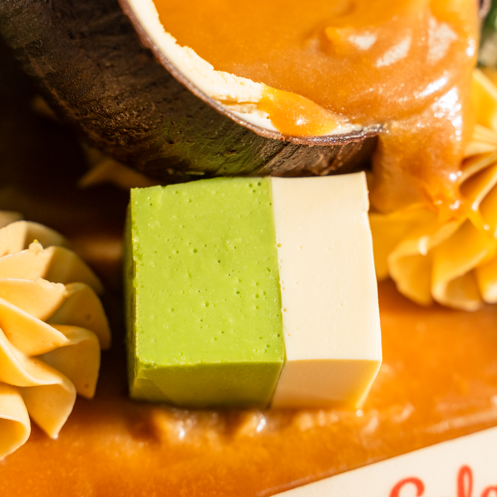 Close-up of a pandan and coconut kuih talam cube on the Muhibbah cake, beside a coconut shell of gula melaka sauce