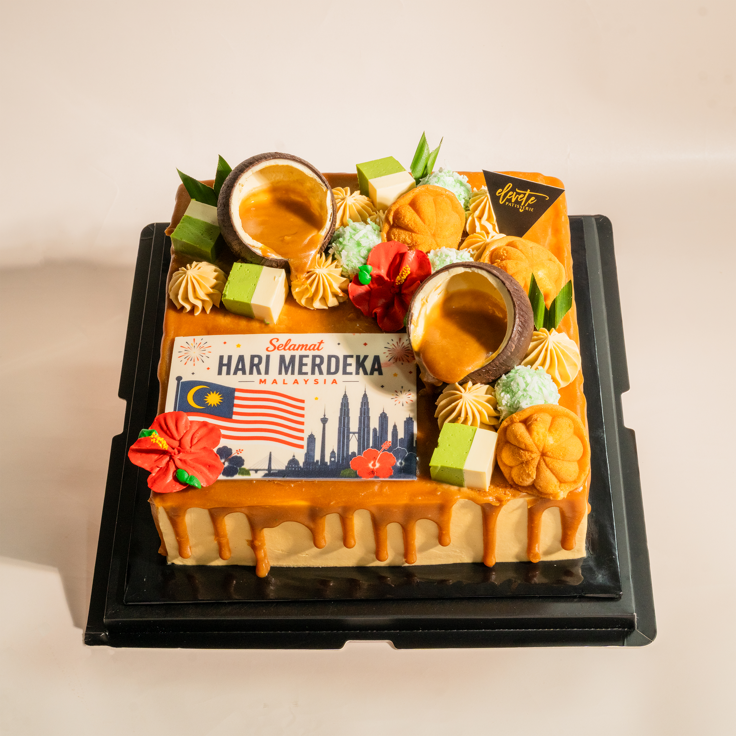 Muhibbah Merdeka kuih cake from above — Selamat Hari Merdeka plaque with Malaysian flag and KL skyline, coconut shells of gula melaka, kuih talam cubes, kuih bahulu and ondeh-ondeh truffles